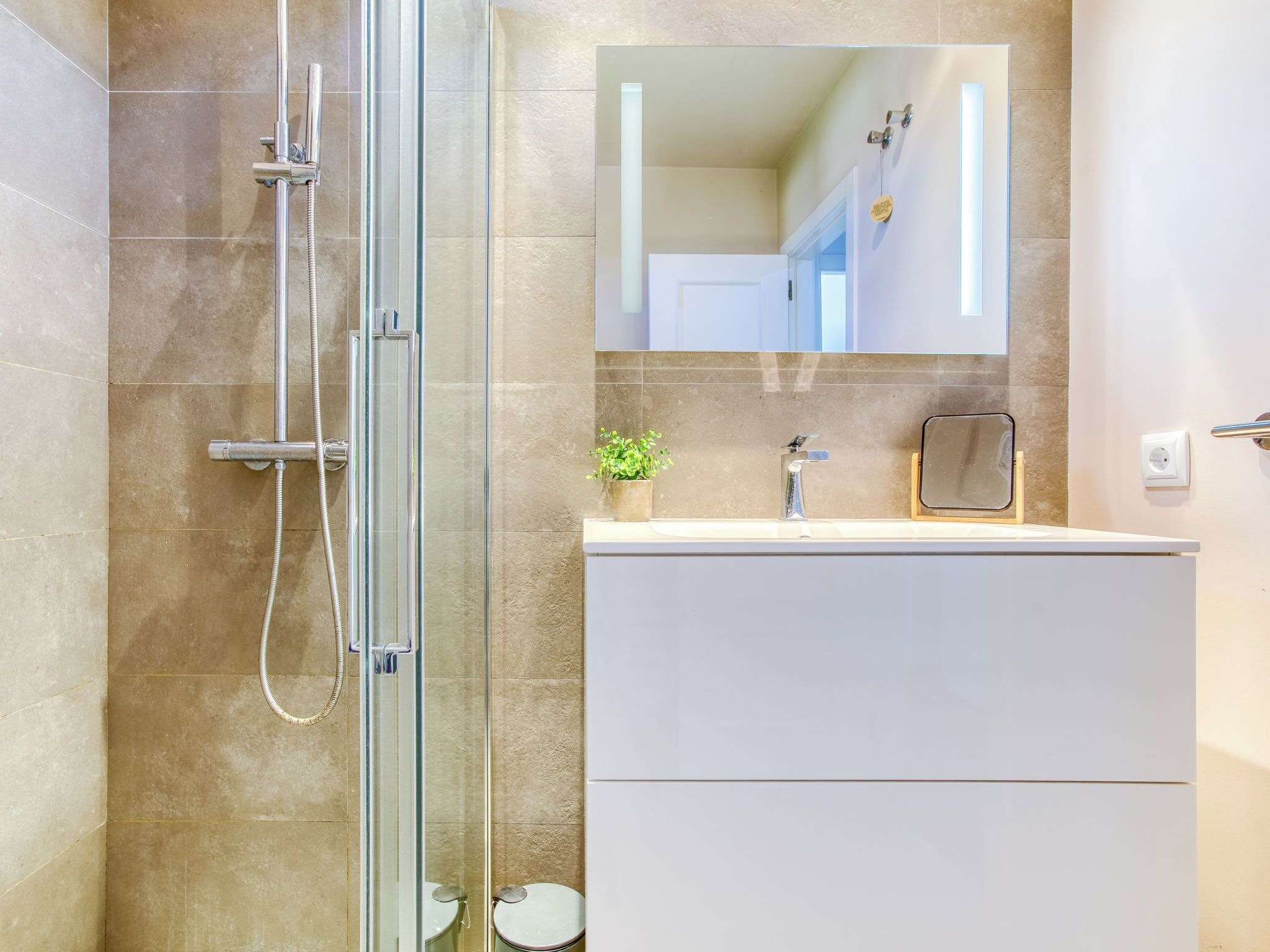 Un baño con cabina de ducha de vidrio, un tocador blanco con espejo y una pequeña planta en maceta.