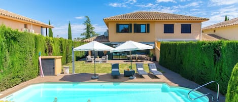 A swimming pool with lounge chairs and umbrellas, surrounded by a well-maintained hedge and a building with a tiled roof.
