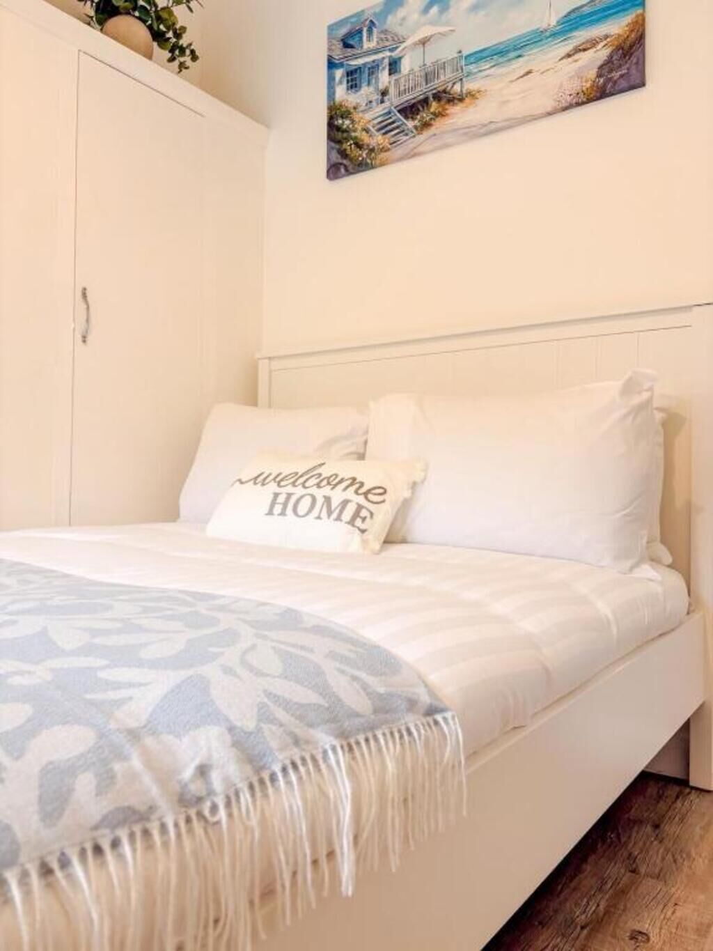 A bedroom with a bed, a welcome home pillow, a painting of a beach house, and a wardrobe.