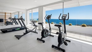 A gym with treadmills and an elliptical machine, large windows with a view of the ocean, and a modern interior design. - Blue Infinity I by Interhome (Moraira)