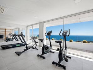 Ein Fitnessraum mit Laufbändern und einem Crosstrainer, großen Fenstern mit Meerblick und modernem Design.
