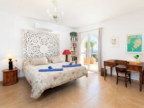 Une chambre à coucher comprenant un lit, une table de chevet, une lampe, une bibliothèque, un bureau, une chaise et une vue sur l’extérieur, visible à travers une porte vitrée.