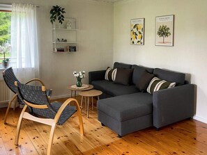 A living room with a grey sectional sofa, a wooden chair, a small round table with a vase, and a white shelving unit with decorative items. - 4 Person Holiday Home in Hacksvik (Håcksvik)