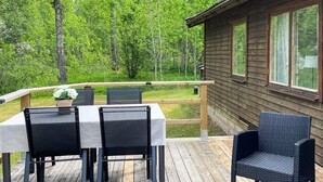 Een houten terras met een tafel en stoelen, een rieten stoel, en een hutje op de achtergrond.