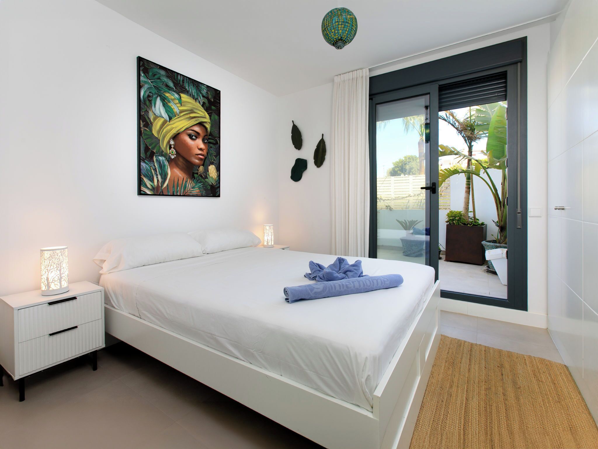 A modern bedroom with a bed, bedside tables, a painting, and a view of the outdoors.