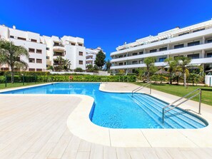 A swimming pool with a wooden deck, surrounded by a well-maintained lawn and palm trees, with multi-story buildings in the background. - Sea Garden by Interhome (Torrox Costa)