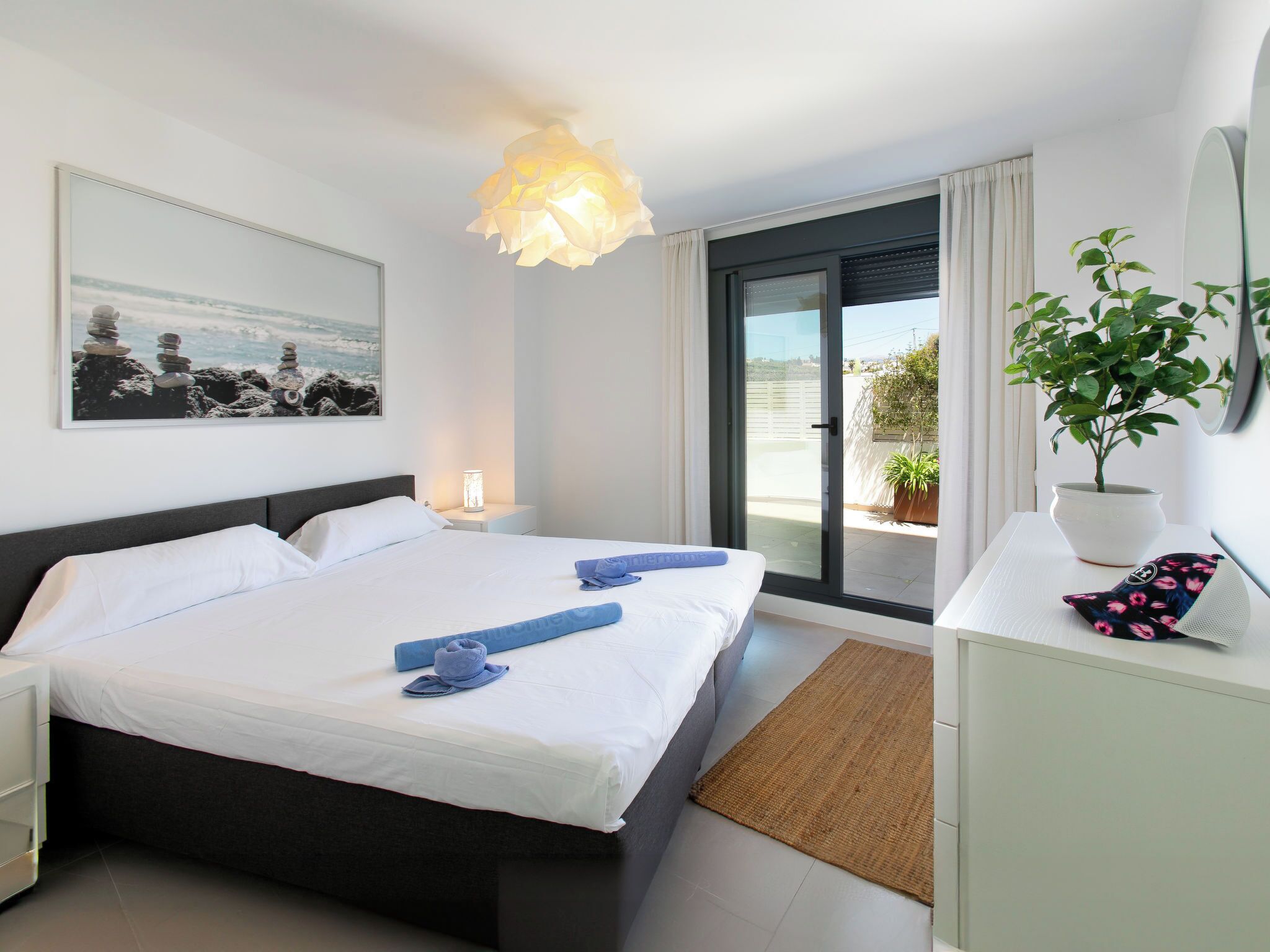 Une chambre d’hôtel comprenant un lit, une table de chevet, une commode, une lampe, une fenêtre avec vue, une plante en pot et un tableau au mur.