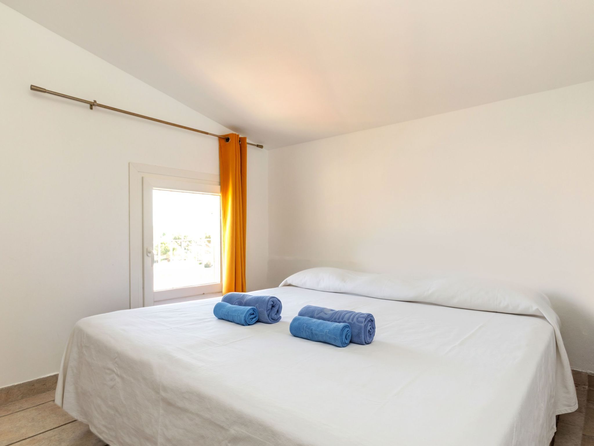 A bedroom with a bed, two rolled towels, a window with curtains, and a door.