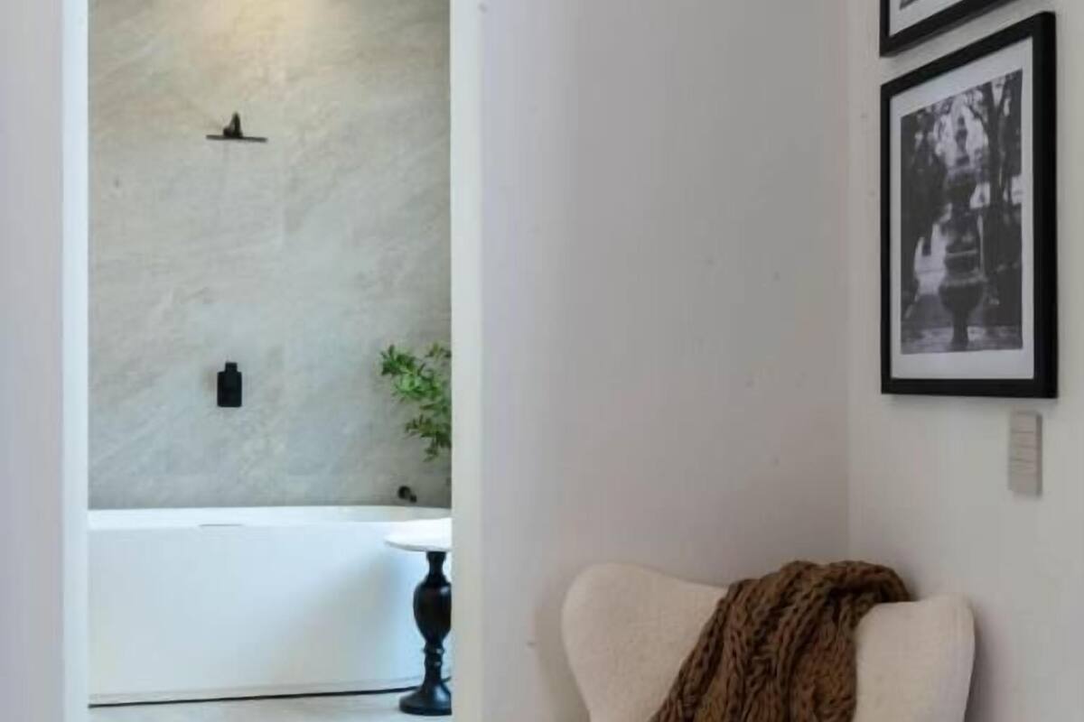 Habitación de esquina con un sillón blanco, una mesita redonda y un cuadro enmarcado en la pared.