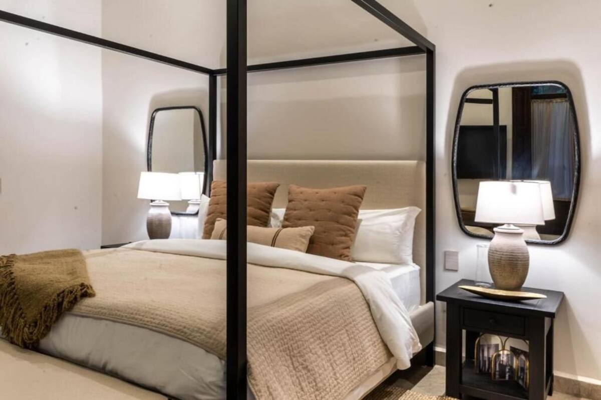 Una cama con dosel, con un edredón y almohadas color beige, una mesita de noche con una lámpara y un espejo en la pared.