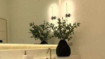 Un baño moderno con una encimera blanca, un florero negro con plantas, un espejo y un perchero para toallas.