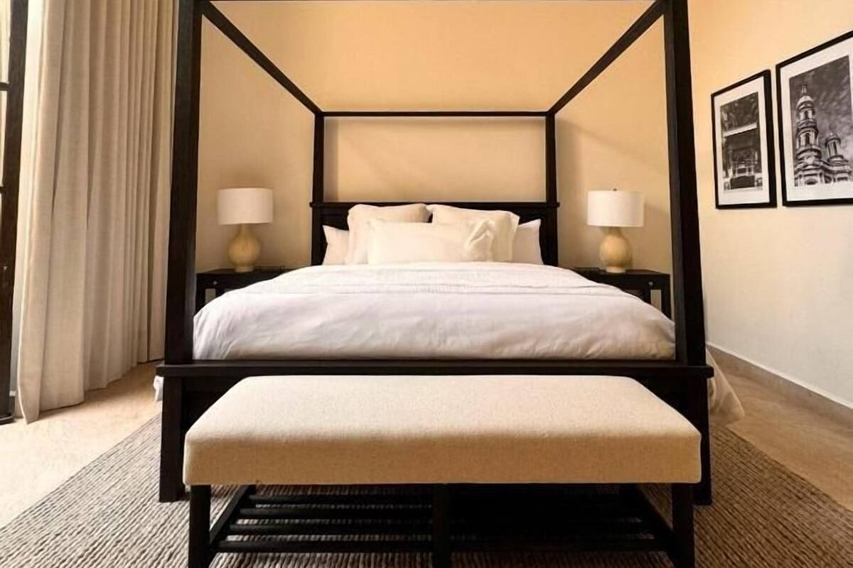 Una cama con dosel, un banco a juego, dos mesitas de noche con lámparas, una alfombra con textura y un aire acondicionado instalado en la pared.