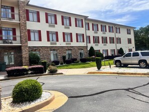 A multi-story hotel with a stone facade, red shutters, and a parking area with a white SUV. - Haton Suites (Conyers)