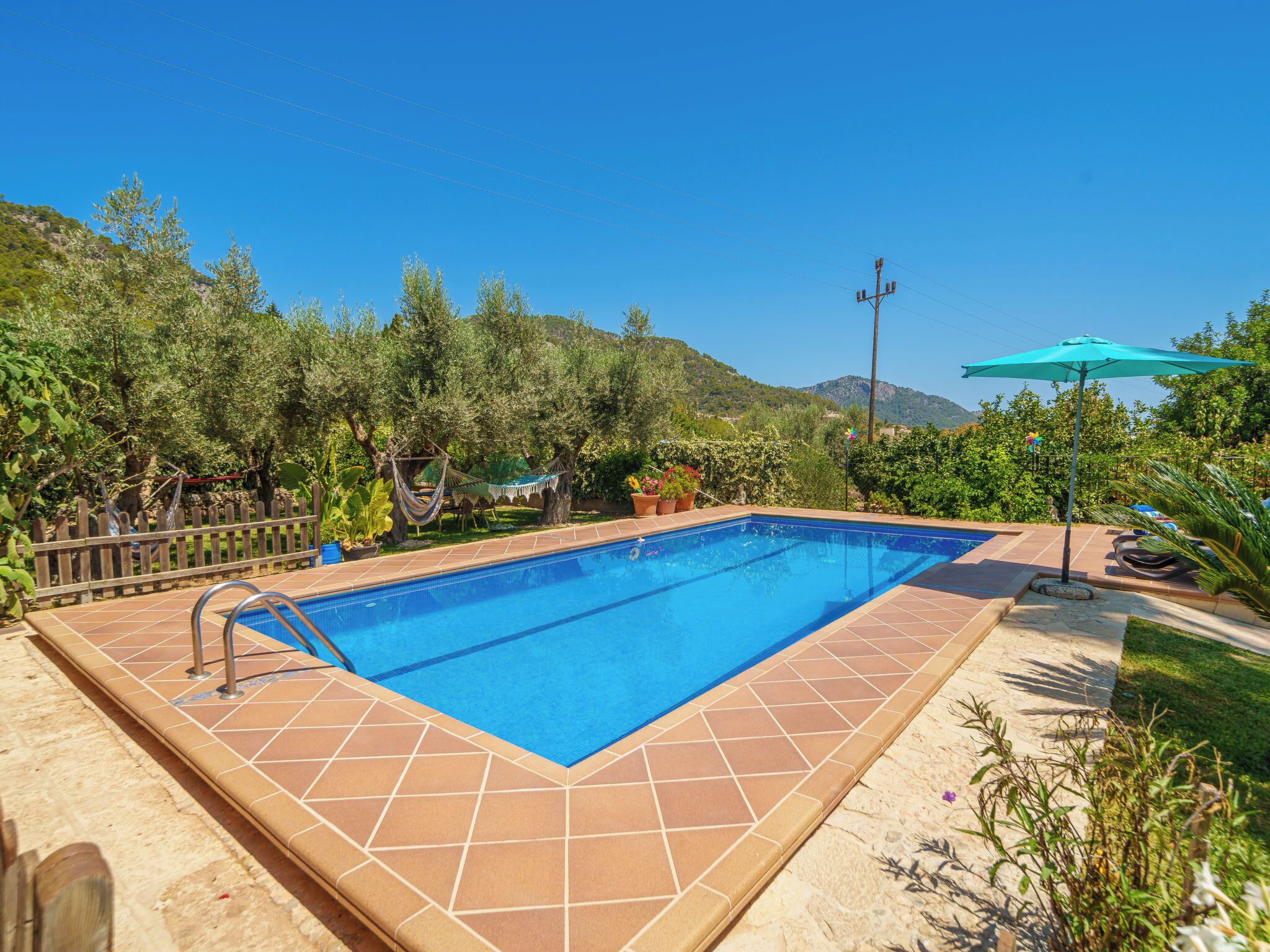 A rectangular swimming pool with a tiled border, surrounded by a wooden fence, potted plants, and a hammock.