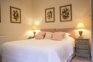 Ein ordentlich bezogenes Bett mit weißen Leinen, zwei Nachttischlampen und drei gerahmte botanische Drucke an der Wand.