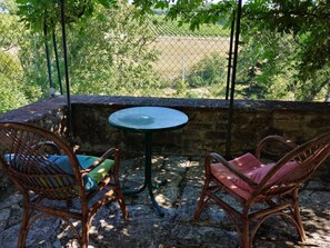 Eine Terrasse mit zwei Korbstühlen, einem kleinen runden Tisch und Blick auf ein eingezäuntes Feld durch ein Maschendrahtgitter.