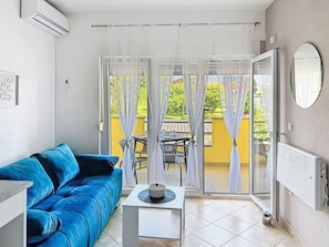 A room with a blue sofa, a small table with a hat, a radiator, and a view through a glass door to an outdoor area with a table and chairs. - Rondine by Interhome (Novigrad (Istra))
