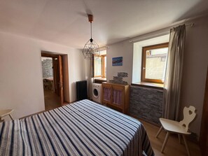 Een slaapkamer met een bed, een wasmachine, een radiator, een raam met gordijnen en een stoel.