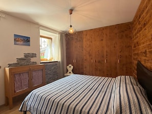 A bedroom with a bed, wooden wardrobe, a small table with a checkered cloth, a wall-mounted shelf, a window with curtains, and a hanging light fixture. - Casa Le Pale by Interhome (Fiera di Primiero)