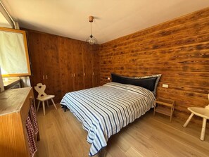 A bedroom with a wooden wall, a bed with a striped bedspread, a chair, a desk, and a hanging light fixture. - Casa Le Pale by Interhome (Fiera di Primiero)