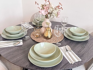 Ein runder Esstisch mit grünen Tellern und klaren Gläsern, eine Blumenvase und eine gewebte Tischmatte.