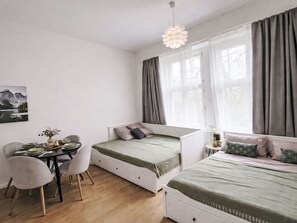 Ein Zimmer mit Esstisch, zwei Betten, einem Fenster mit Vorhängen und einer Pendelleuchte.