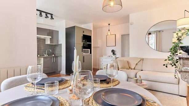 Uma área de refeições com uma mesa posta para quatro pessoas, um sofá, um espelho e uma cozinha ao fundo.