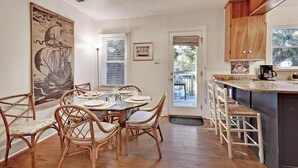 A dining area with a round table, wicker chairs, a large painting, and a kitchen with wooden cabinets and a coffee station.