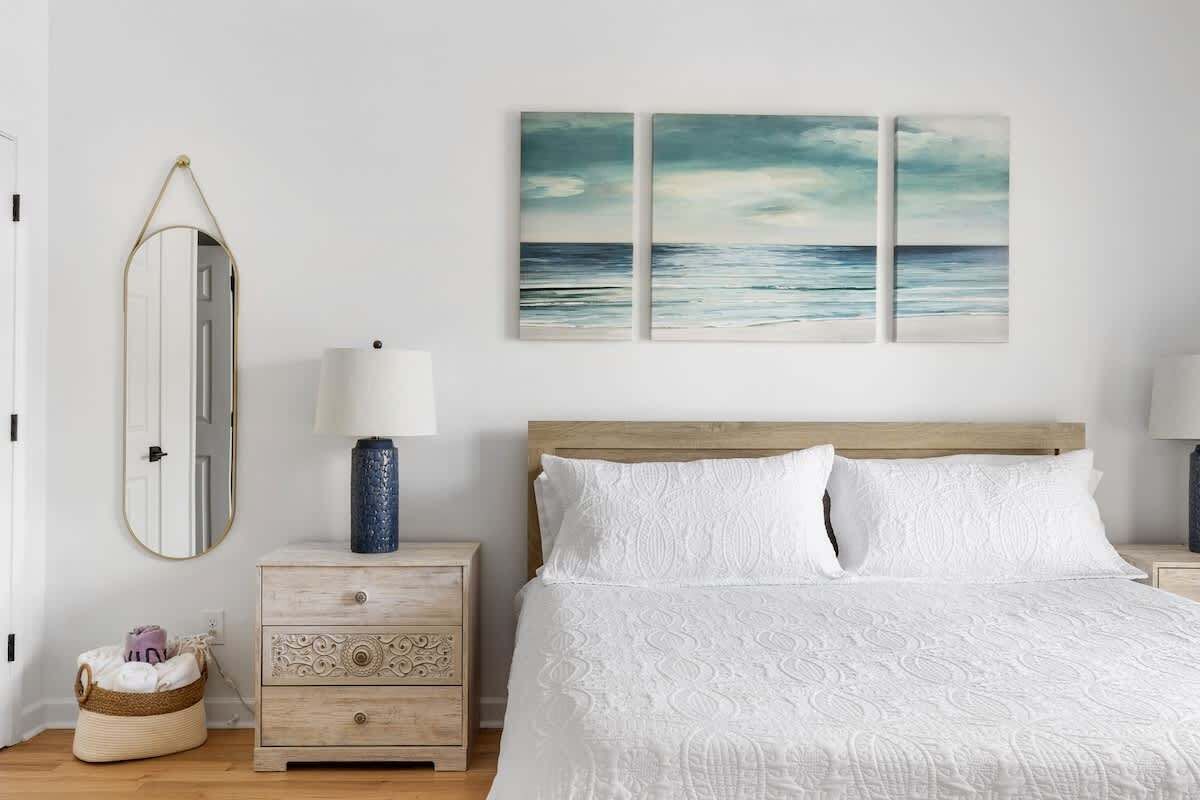 Een slaapkamer met een houten hoofdbord, een wit sprei, een nachtkastje met een lamp, een spiegel en twee aan de muur hangende schilderijen.