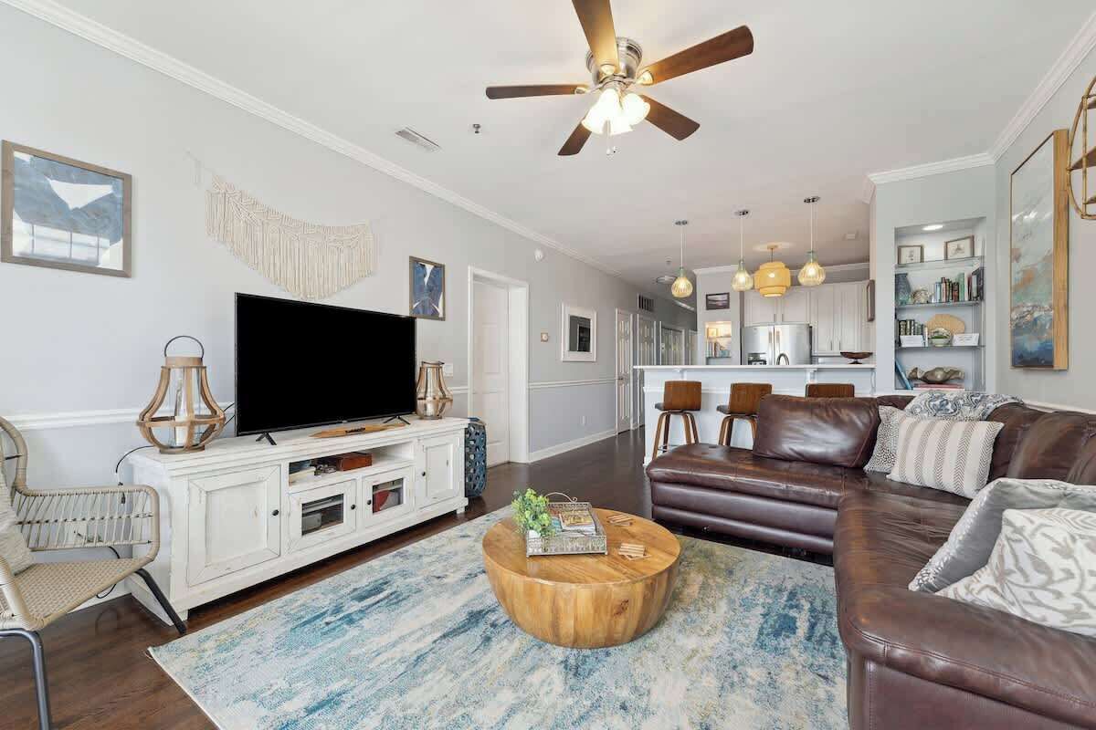 A living room with a television, a ceiling fan, a brown leather sofa, a round coffee table, and a kitchen in the background.