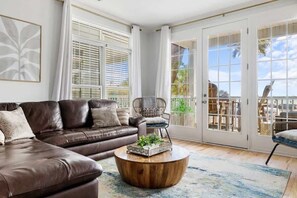 Ein Wohnzimmer mit braunem Ledersofa, einem runden Holztisch, einem Korbstuhl und einer großen Schiebeglastür, die zu einem Außenbereich führt.