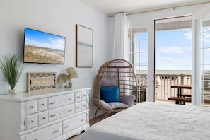 Ein Schlafzimmer mit einem Bett, einem Schminktisch, einem Korbstuhl, einem Fernseher und einem Balkon mit Blick.