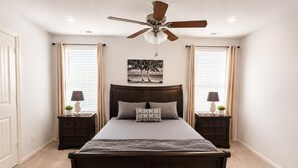 Un dormitorio con una cama grande, un ventilador de techo, dos mesitas de noche con lámparas, un cuadro en la pared y una ventana con cortinas.