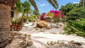 A tropical garden with palm trees, a rock formation, and a small building with outdoor seating. - Villa Nirvana 6BR Private Family, Pool & Bar Villa (Noord)