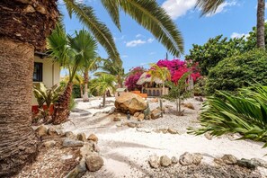 A tropical garden with palm trees, a rock formation, and a small building with outdoor seating. - Villa Nirvana 6BR Private Family, Pool & Bar Villa (Noord)