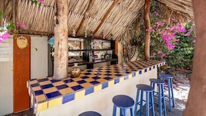 A bar area with a checkered counter, stools, and a thatched roof. - Villa Nirvana 6BR Private Family, Pool & Bar Villa (Noord)