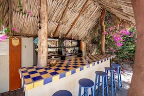 A bar area with a checkered counter, stools, and a thatched roof. - Villa Nirvana 6BR Private Family, Pool & Bar Villa (Noord)