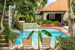 A pool with lounge chairs, surrounded by tropical plants and a house with a tiled roof. - Villa Nirvana 6BR Private Family, Pool & Bar Villa (Noord)