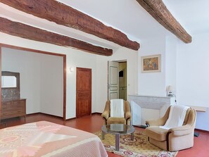 Een kamer met een bed, twee fauteuils, een tafeltje, een houten plafond, een deur en een schilderij aan de muur.