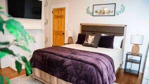 A bedroom with a bed, a TV, a door, a bedside table with a lamp, and a wall decoration. - Safe and quiet street. Walk to shops & more. Fenced in yard w huge front porch.  (Charlotte)