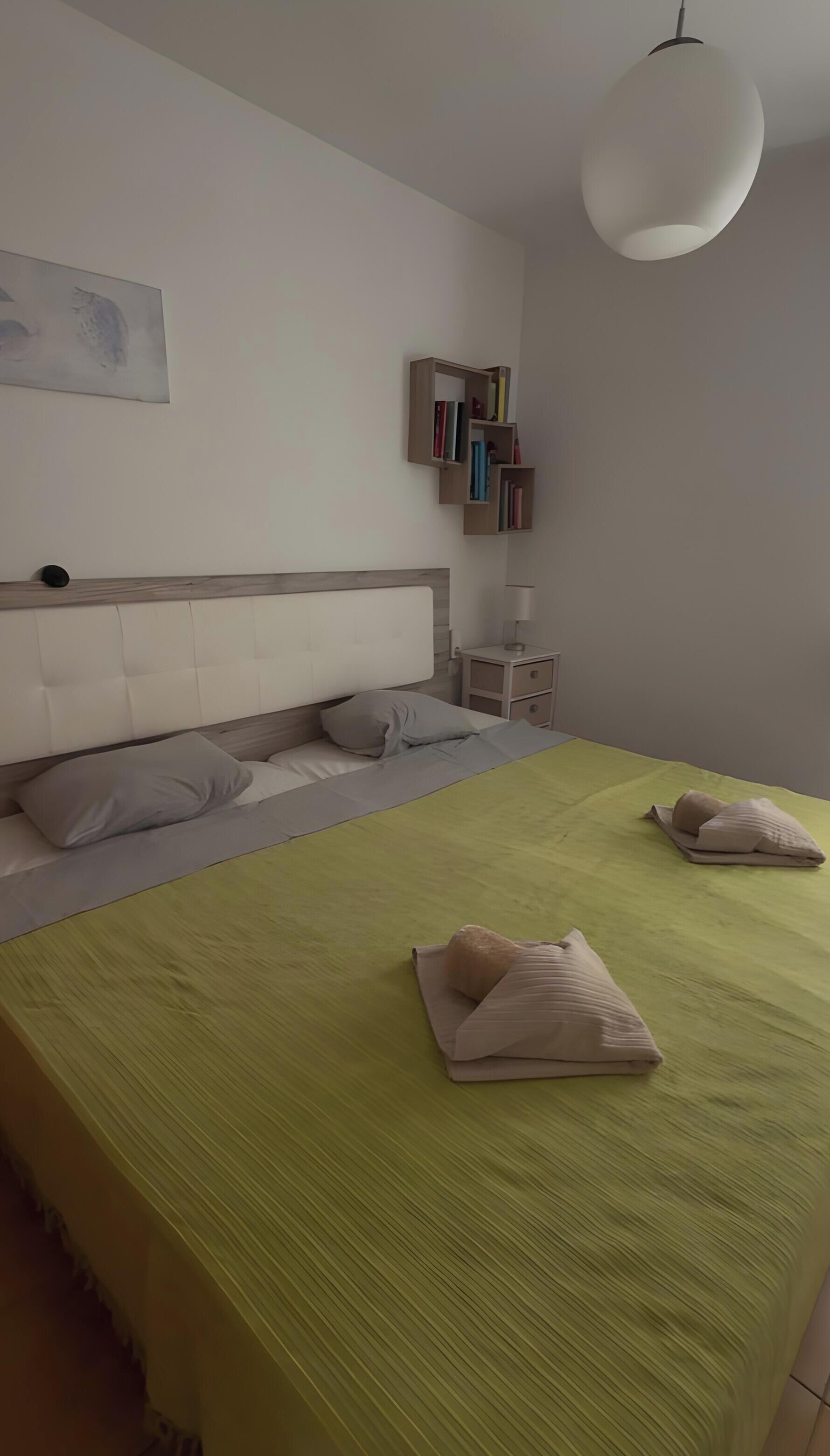 Un dormitorio con cama, almohadas, una mesita de noche, lámpara y estantería en la pared.