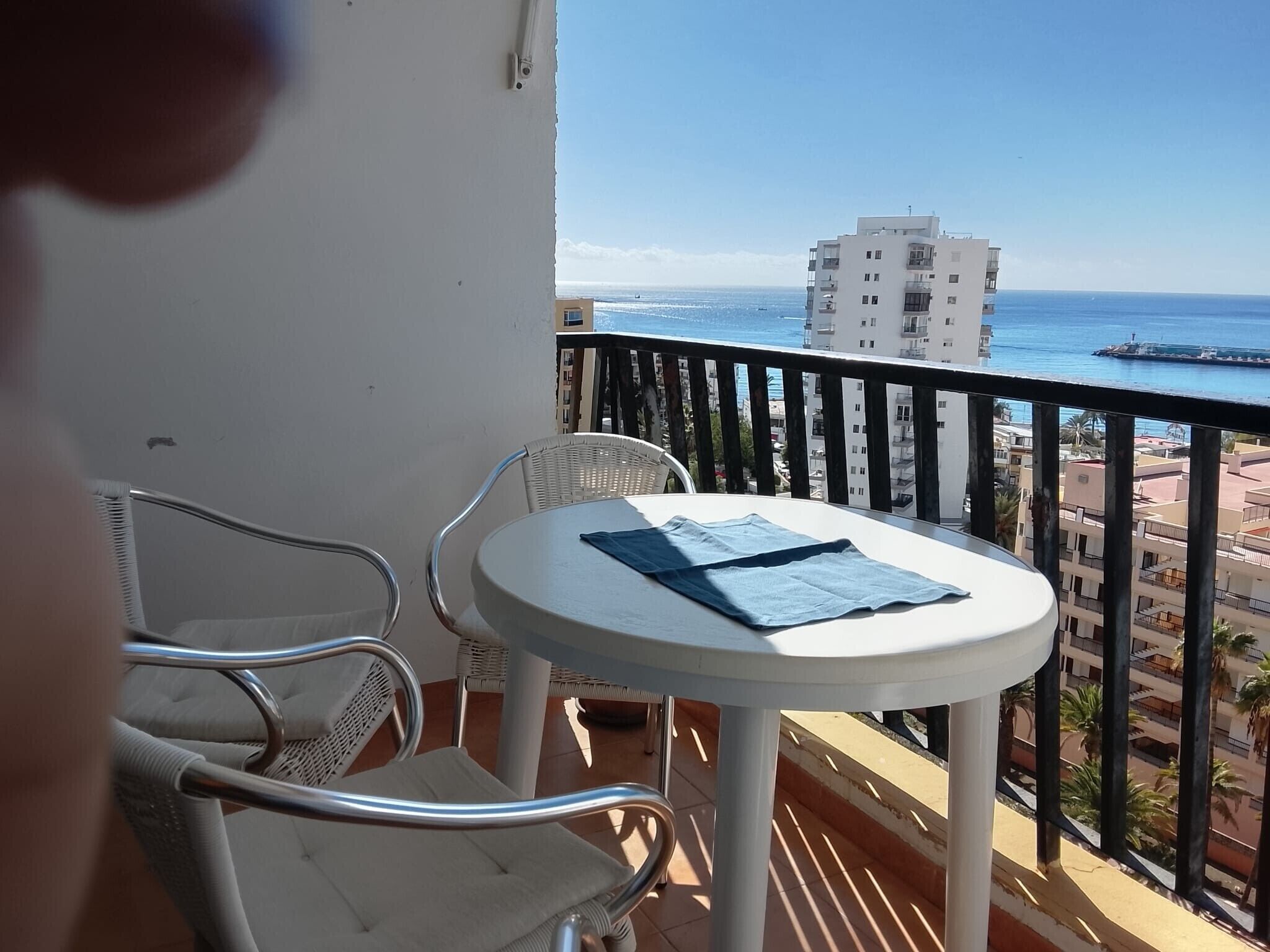 En balkon med et bord og stole, ud til et kystlandskab med bygninger og en bro.