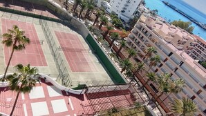A tennis court surrounded by palm trees and buildings, with a view of the sea and a bridge in the background. - 'Achacay Beach' Holiday Apartment with Sea View, Pool and Wi-Fi (Los Cristianos)