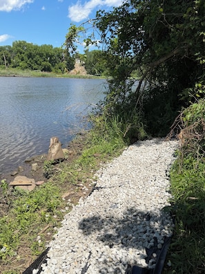 Un sendero de grava junto a un lago tranquilo, con abundante vegetación y árboles al fondo.