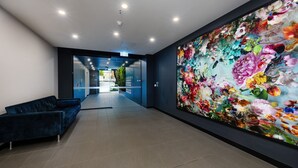 Un vestíbulo moderno con un sofá azul, un amplio mural floral y una pared de vidrio que da a un estacionamiento.