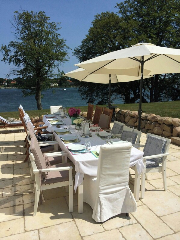 Uma área de refeições ao ar livre com uma mesa posta para quatro pessoas, cercada por cadeiras, perto de um lago com um muro de pedra e árvores.