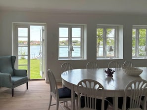 Uma área de refeições com uma mesa redonda, cadeiras brancas e vista para a água através de grandes janelas.