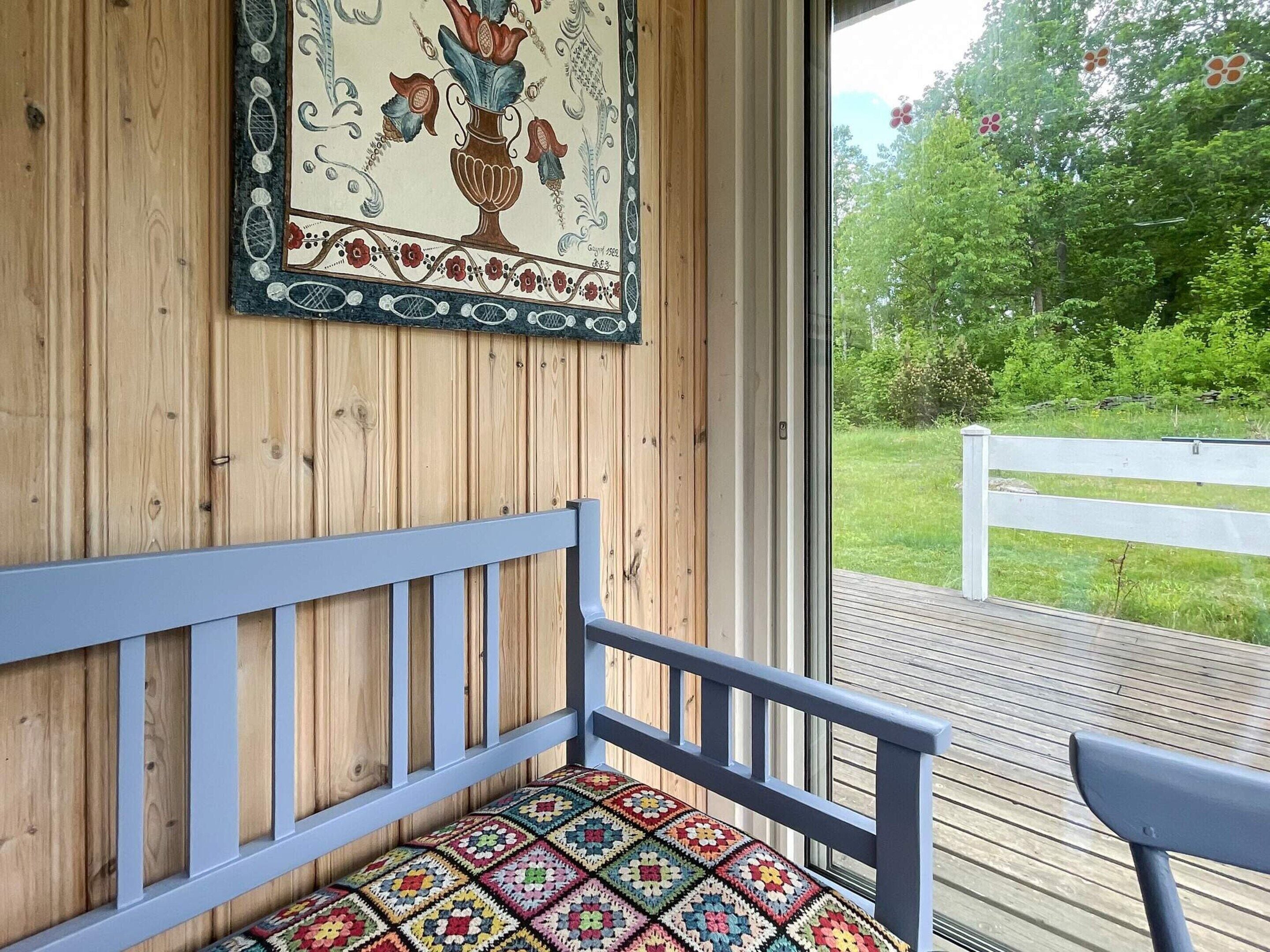 A wooden bench with a colorful cushion, a wall-mounted decorative piece, and a view of a grassy area through a glass door.