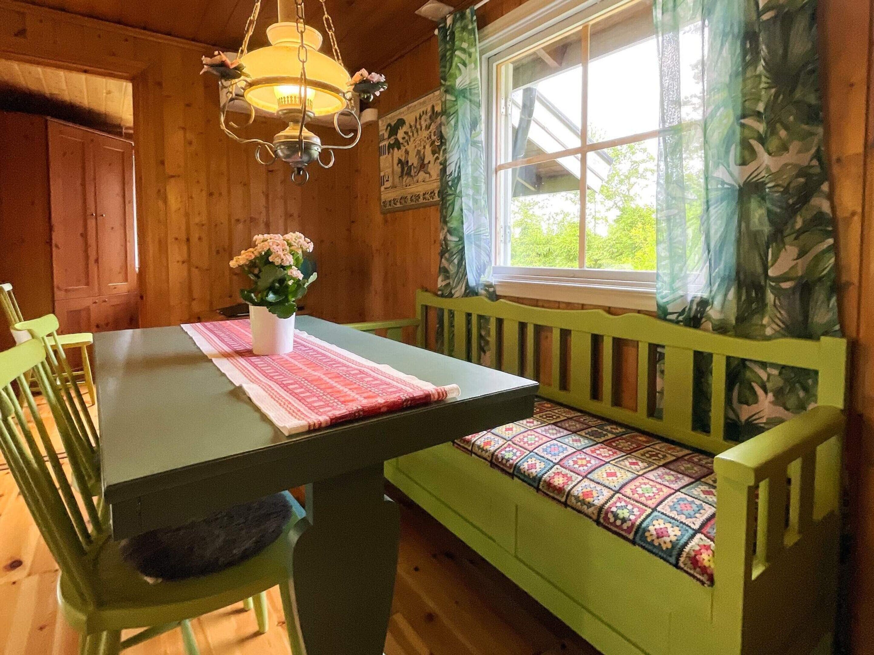 A wooden cabin interior with a dining table, chairs, a bench with a colorful cushion, a hanging light fixture, and a window with curtains.