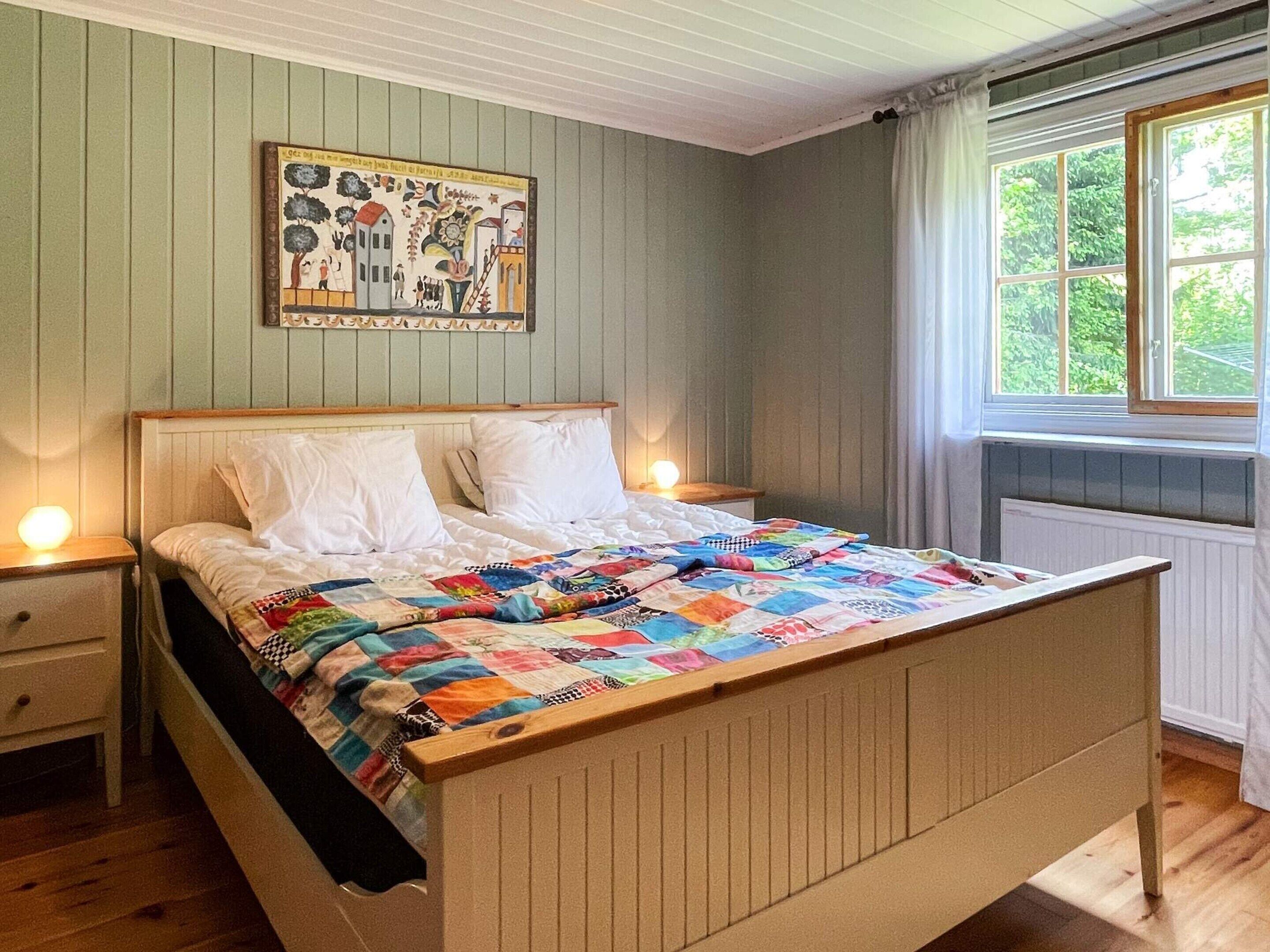A bed with a colorful quilt, a nightstand, a wall painting, and a window with curtains.